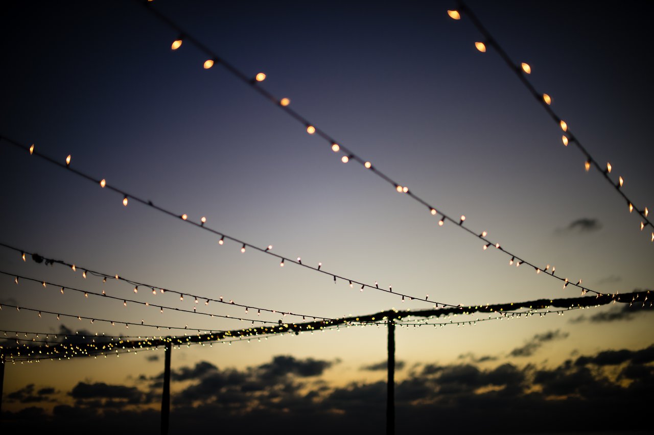 String lights hanging above a beach event at sunset, creating a festive atmosphere for the gathering.