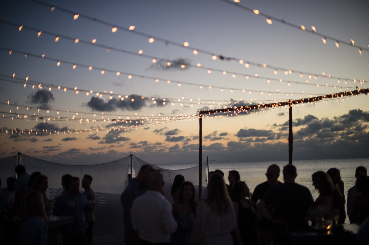 People socializing under string lights at a beachside event during sunset.