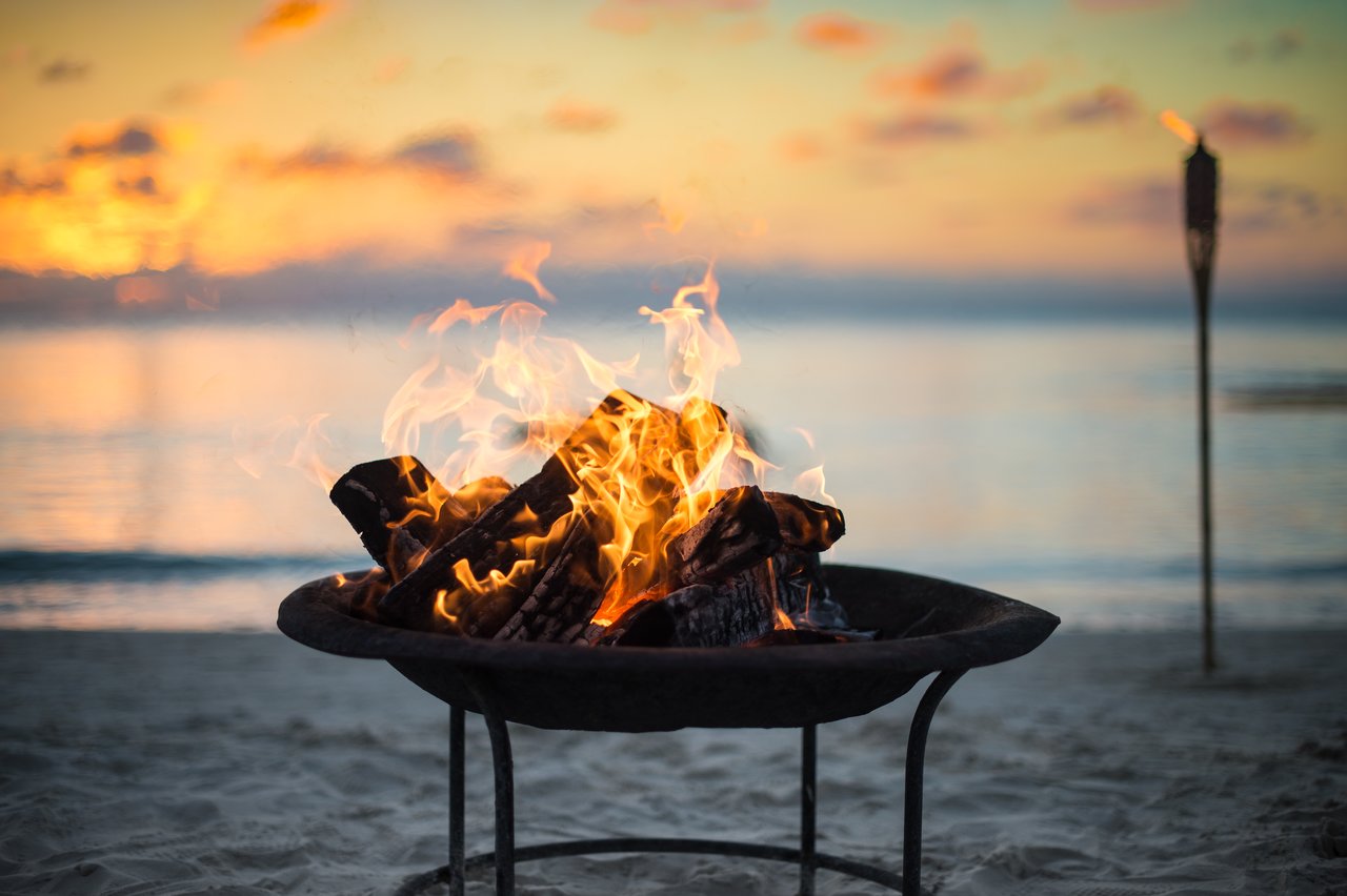 A fire pit with burning wood on a sandy beach at sunset, with the ocean and a tiki torch nearby.