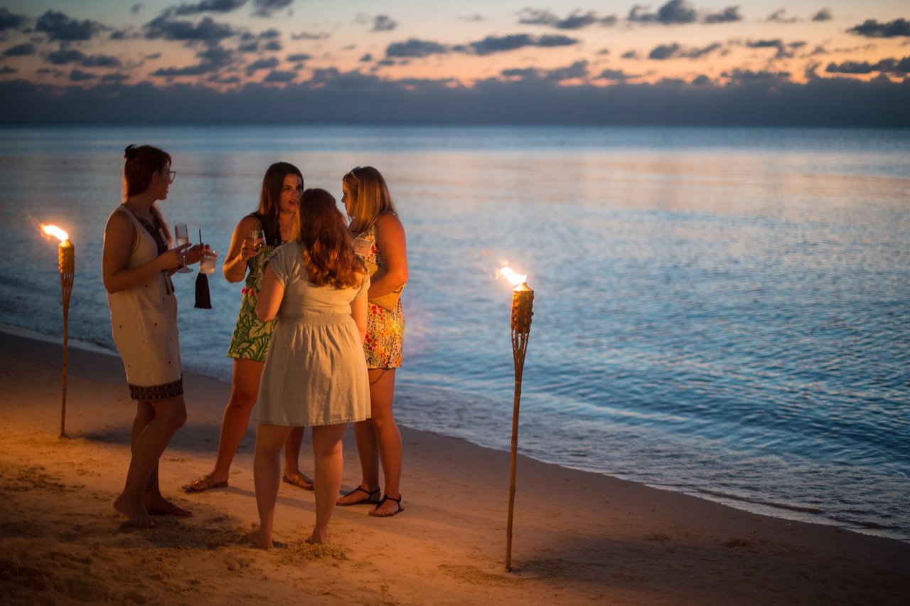 Four women stand on a beach at sunset, holding drinks and talking near lit tiki torches by the water.