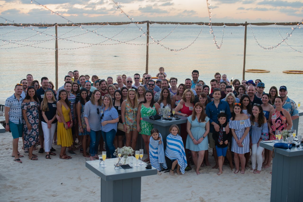 A large group of people poses together on a sandy beach during a company trip.