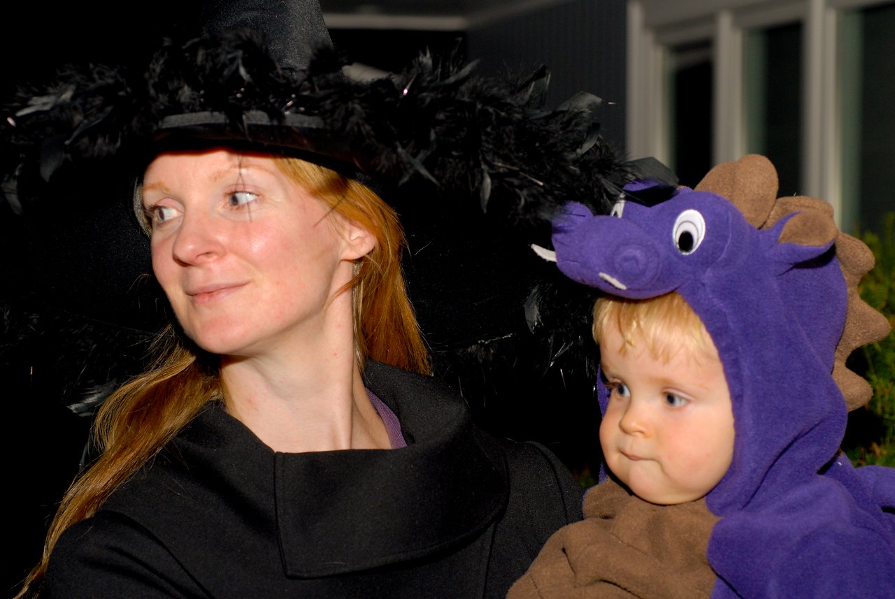 A woman in a witch costume holds a baby dressed as a purple dinosaur during a Halloween event.