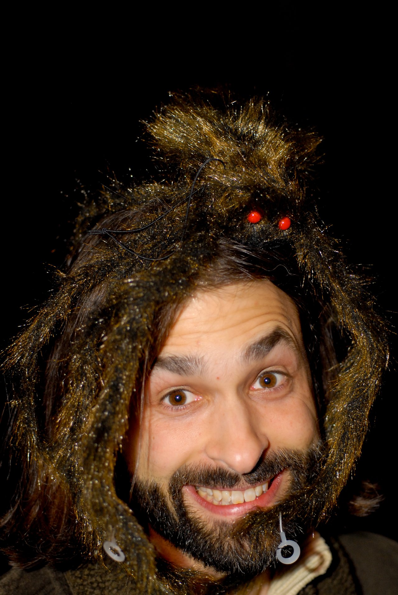 A person smiles while wearing a Halloween costume with fake spider-like hair and red-eyed decorations.