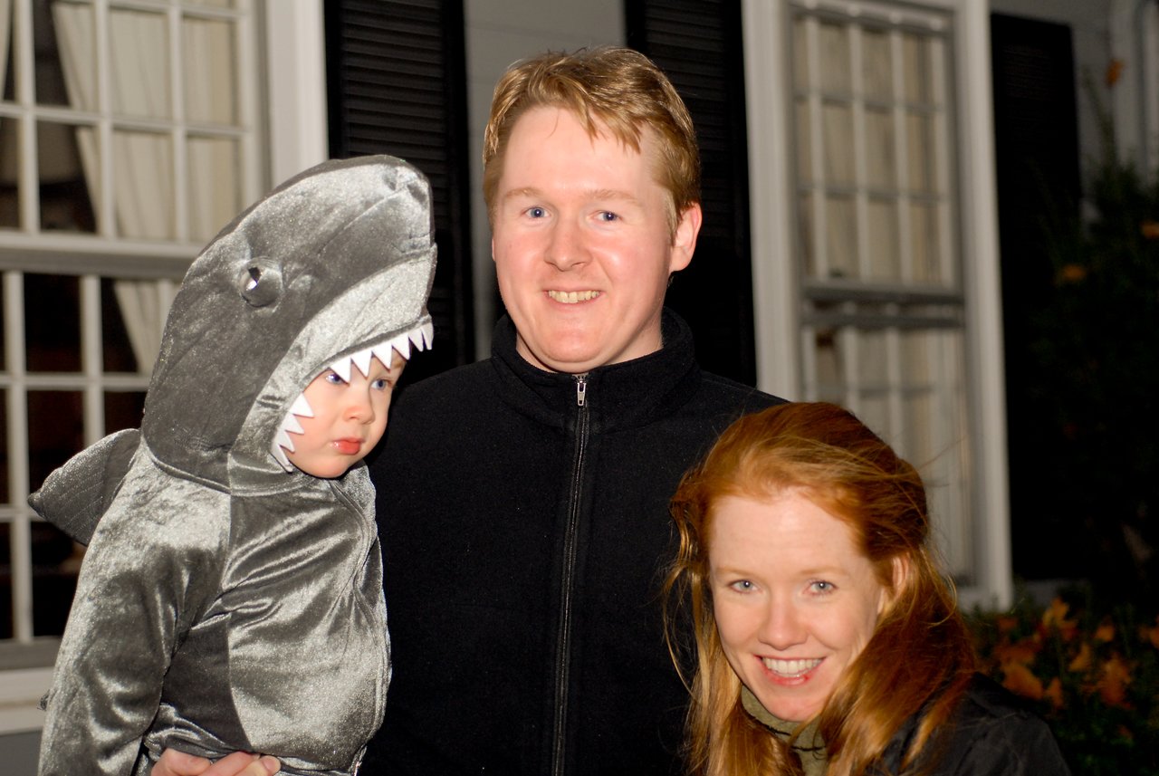 A child in a shark costume is held by an adult, with another person smiling beside them.