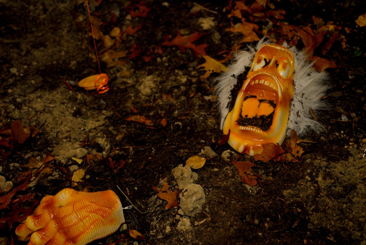 A Halloween decoration of a screaming face and severed hand is partially buried in dirt with scattered leaves.