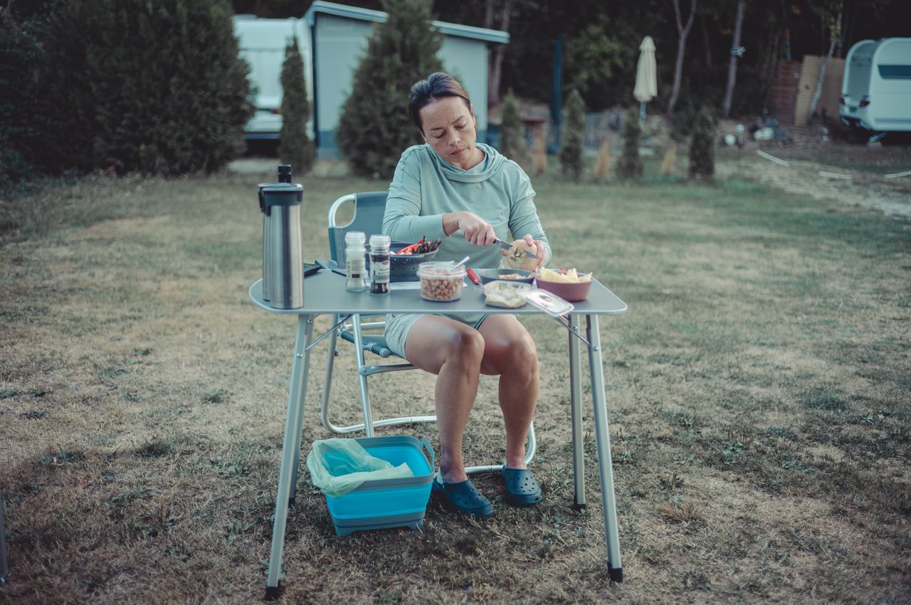 A person sitting at a portable camping table, preparing food with various ingredients and utensils outdoors.