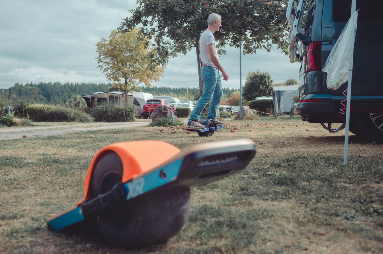 A person rides a Onewheel at a campground, with another Onewheel resting on the grass in the foreground.