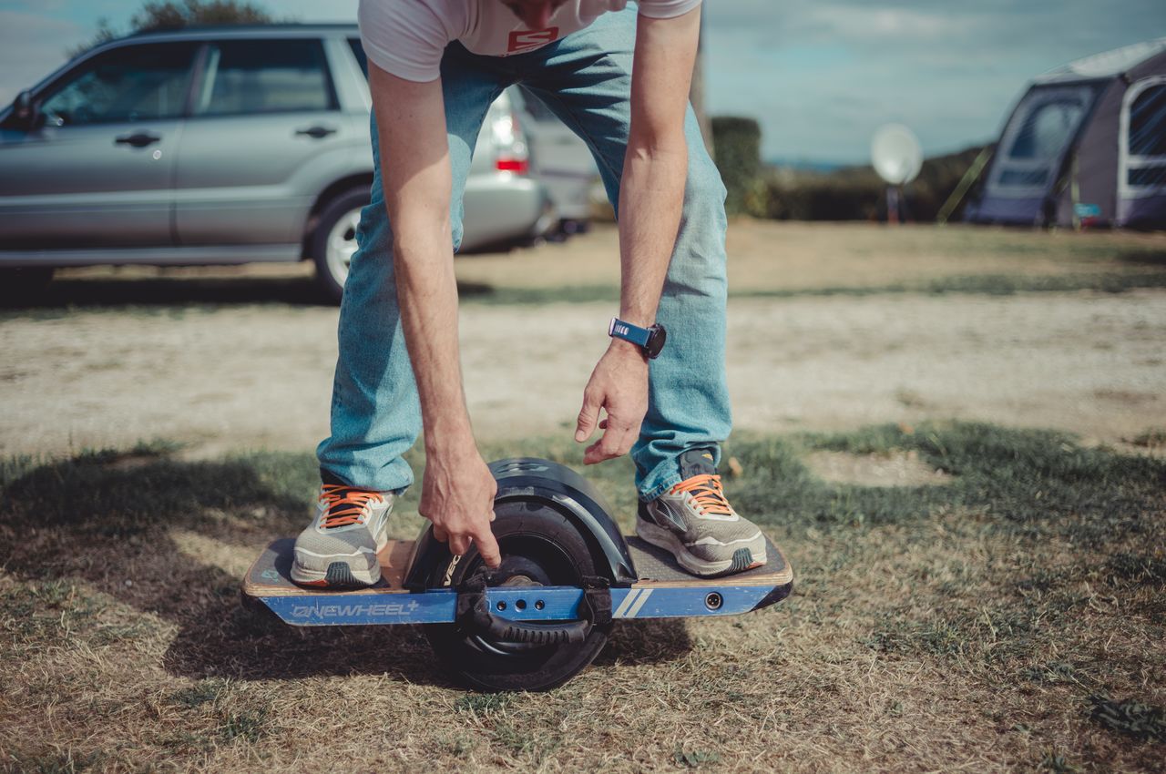 A person rides a Onewheel electric skateboard on grass at a campground, balancing with bent knees and hands down.