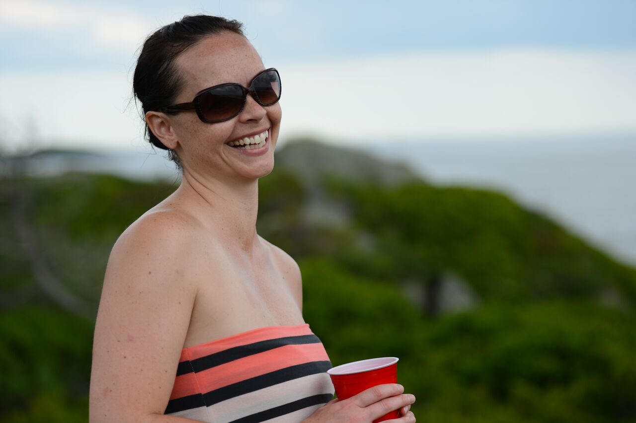 A smiling woman wearing sunglasses holds a red cup outdoors with greenery and the ocean in the background.