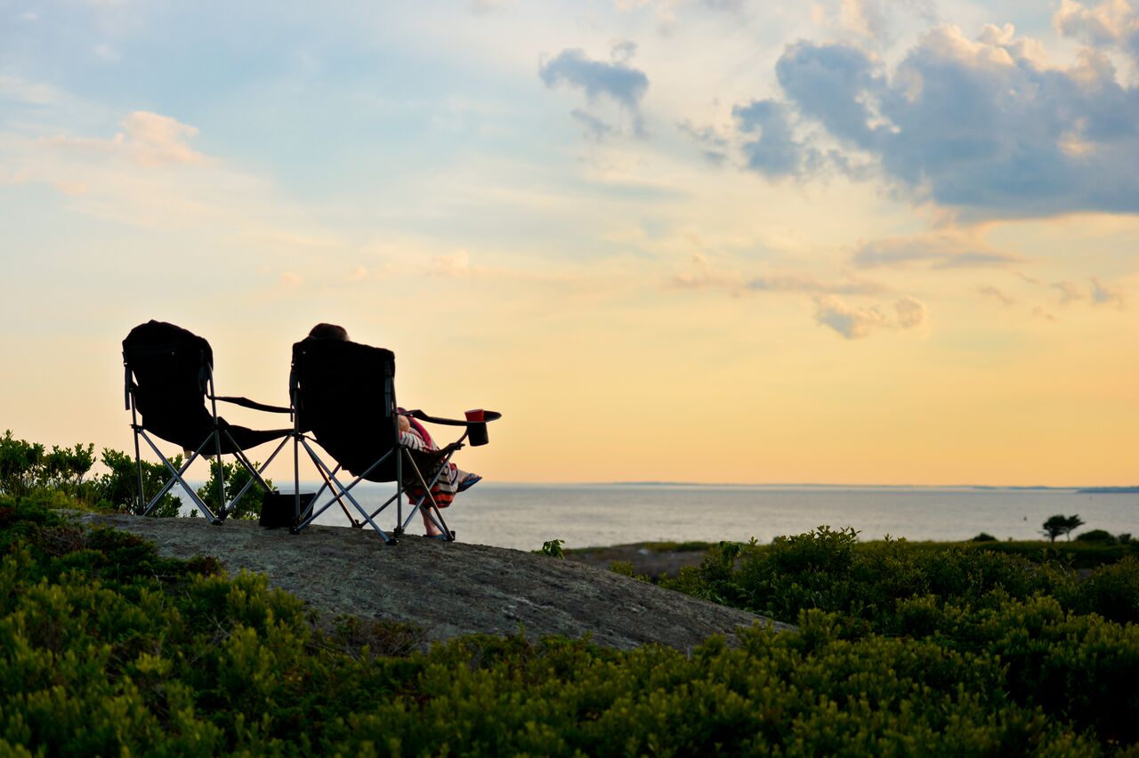 Two camping chairs on a rocky hill overlooking the ocean at sunset, with a person relaxing.