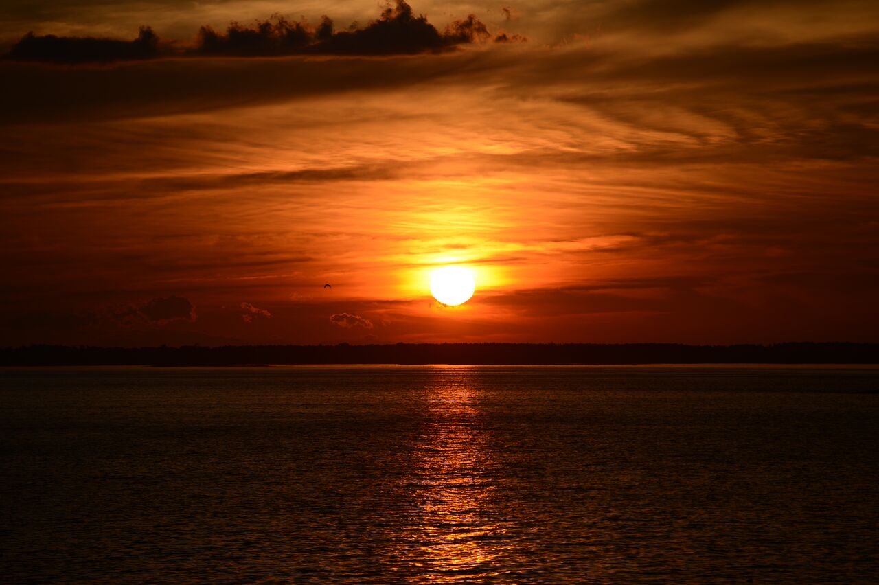 The sun sets over calm water, casting an orange glow across the sky and reflecting on the surface.