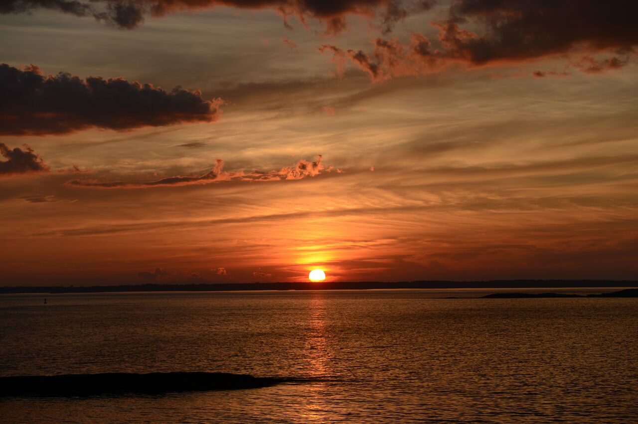 The sun sets over calm ocean waters, casting an orange glow across the sky with scattered clouds.