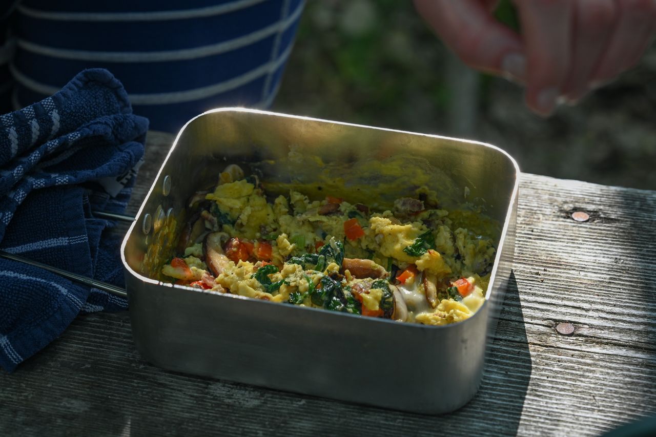 A metal container with scrambled eggs, vegetables, and mushrooms on a wooden picnic table during a camping trip.