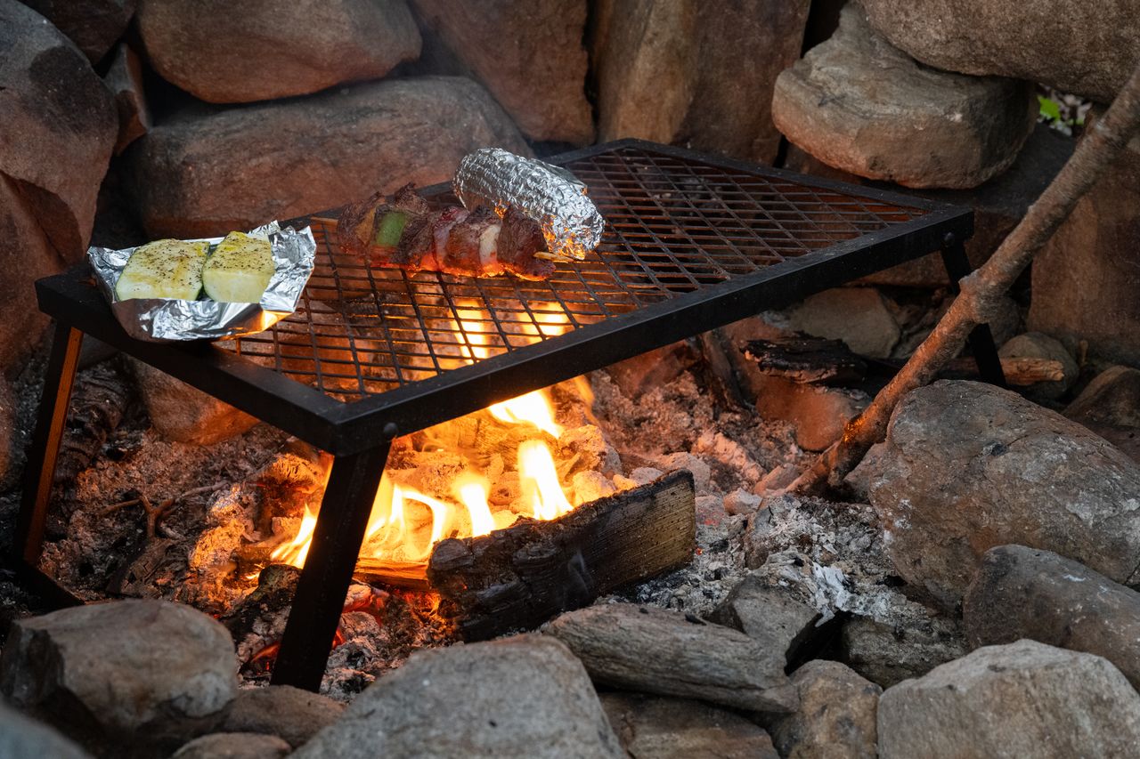 Food cooking over a campfire, including foil-wrapped corn, seasoned zucchini, and skewers with meat and vegetables.