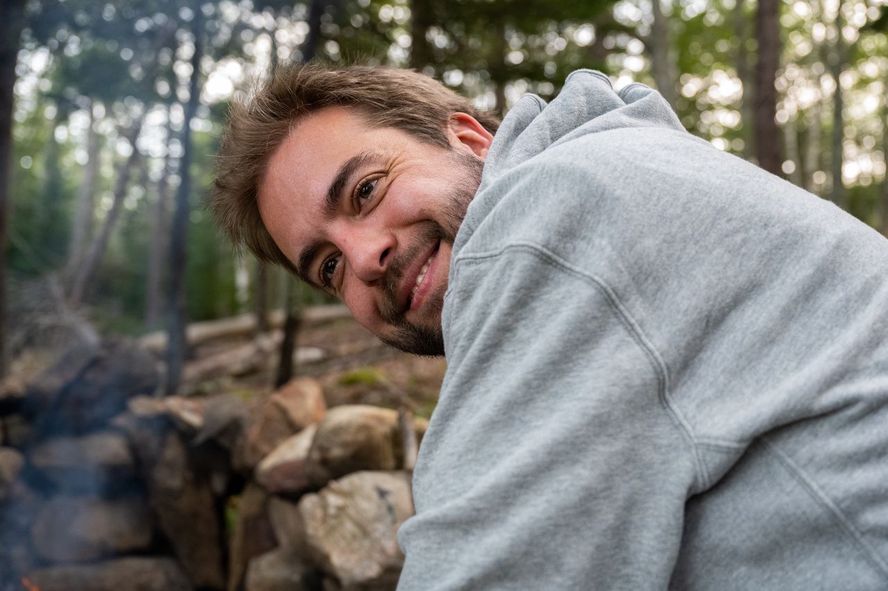 A man in a gray hoodie smiles while tending to a fire at a wooded campsite.