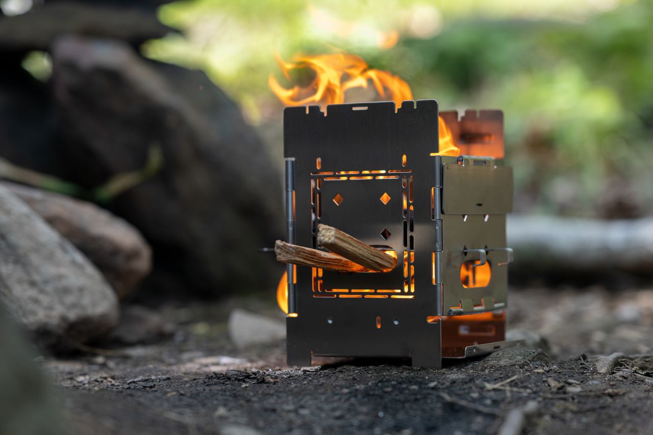 A small metal camping stove with a fire burning inside, fueled by wooden sticks.