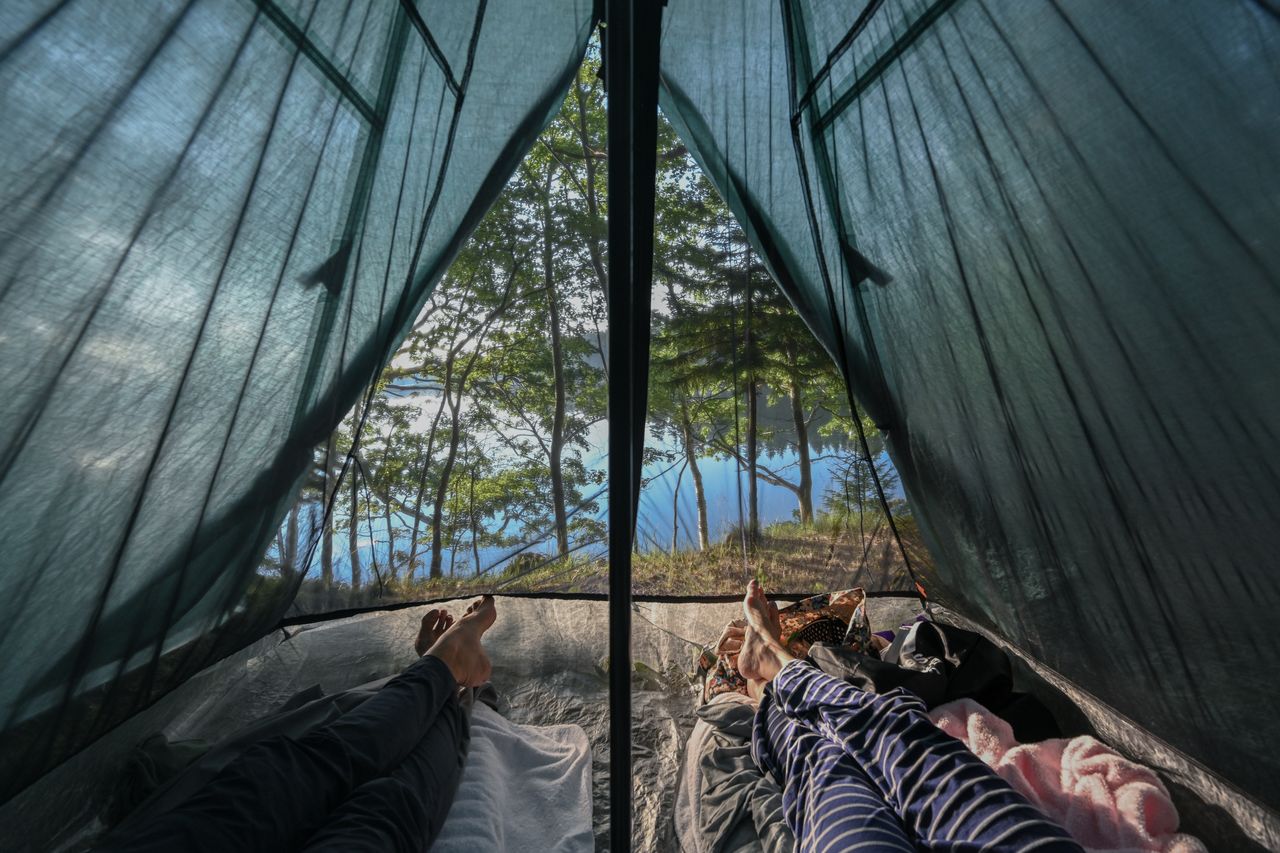Two people lying in a tent, looking out at trees and a lake in the morning.