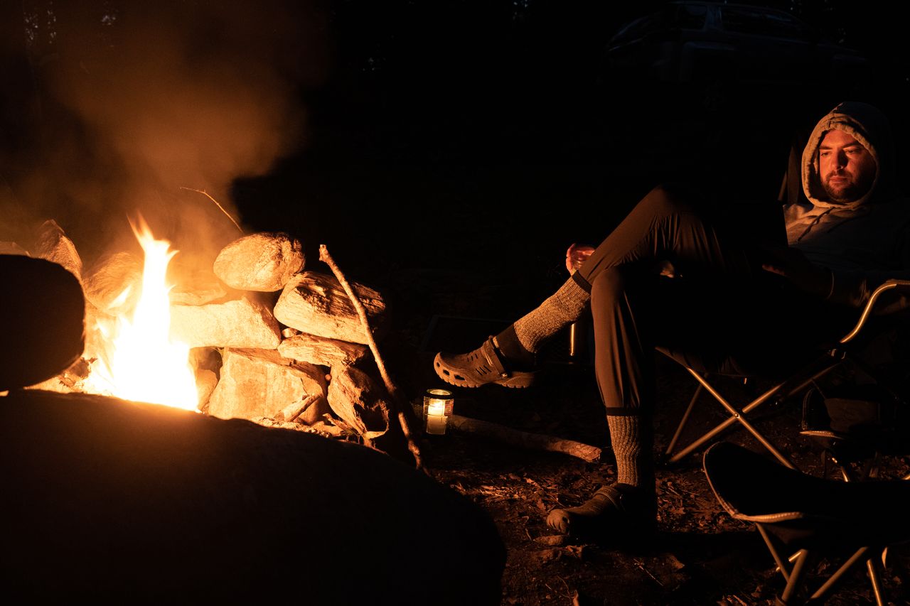 A person sits by a campfire at night, reading and relaxing in a chair.