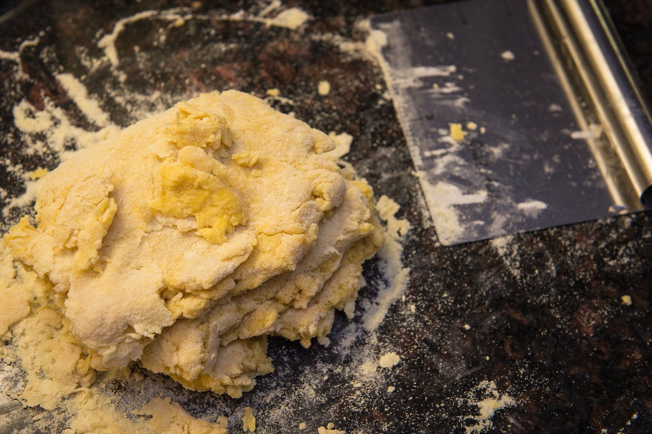 A mound of homemade pasta dough on a floured surface with a metal dough scraper nearby.