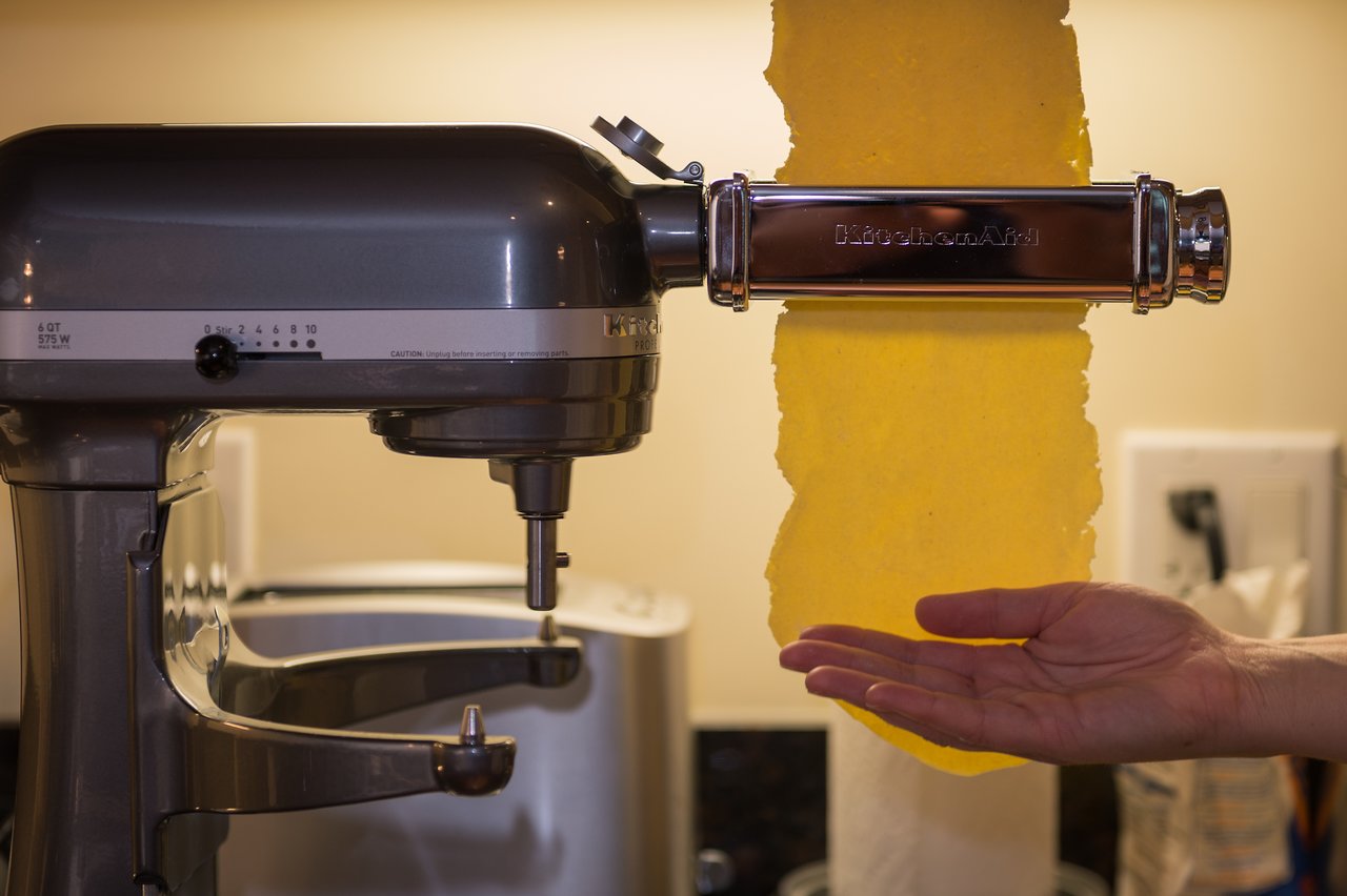 A pasta sheet is being rolled through a stand mixer attachment while a hand supports it underneath.