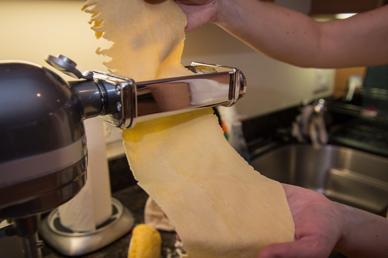A person feeds a sheet of pasta dough through a pasta roller attached to a stand mixer.
