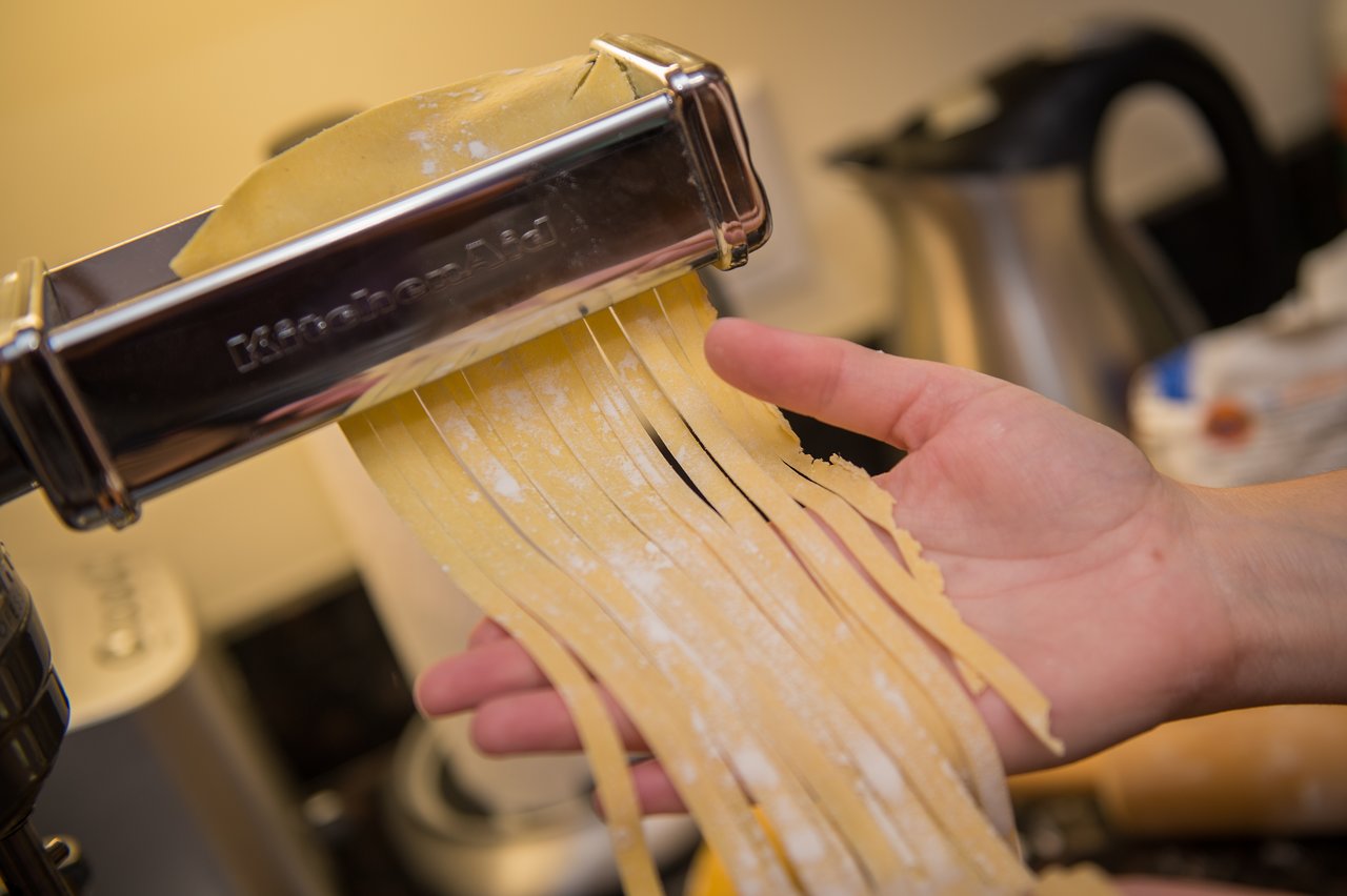 Fresh pasta strips emerging from a pasta machine, guided by a person's hand.