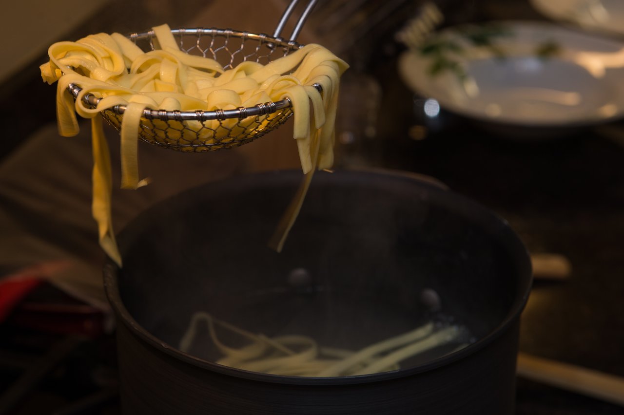 Freshly cooked pasta is lifted from a steaming pot using a metal strainer.