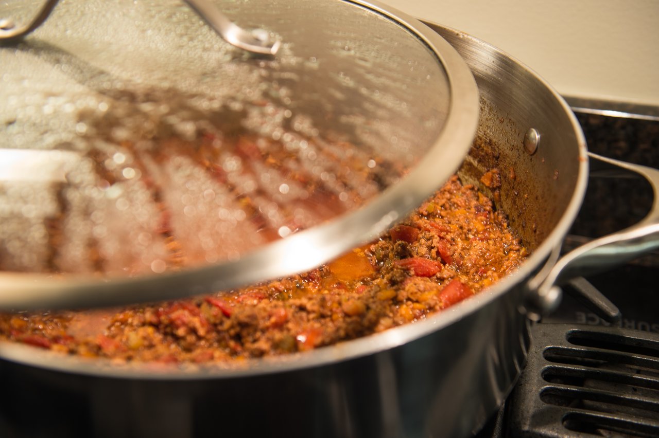 A pot of simmering meat sauce with tomatoes and spices, partially covered by a glass lid on the stove.