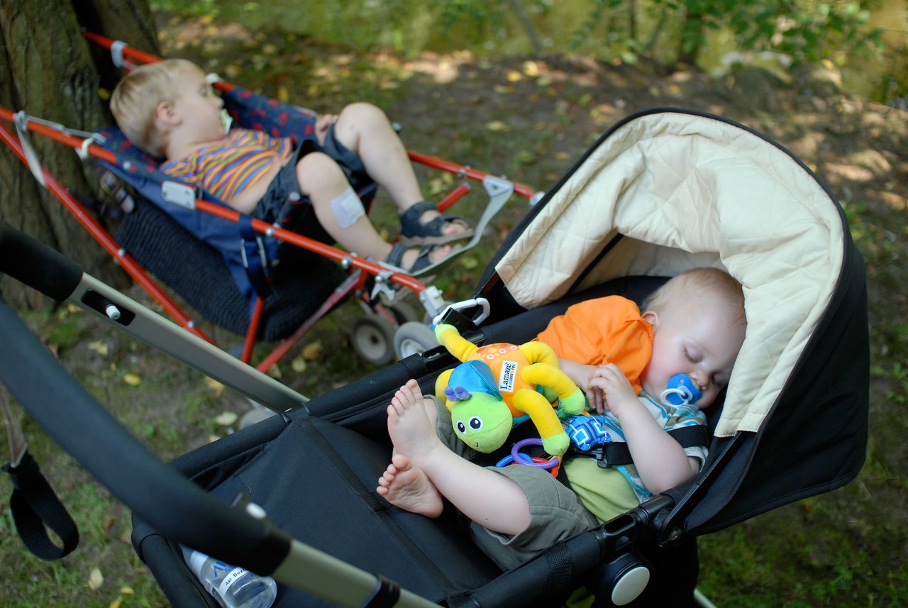 Two young children are sleeping outdoors, one in a stroller and the other in a foldable chair.