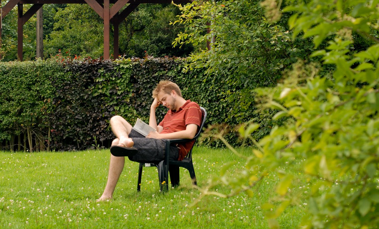 A man sits in a plastic chair on the grass, reading a book with a relaxed posture.