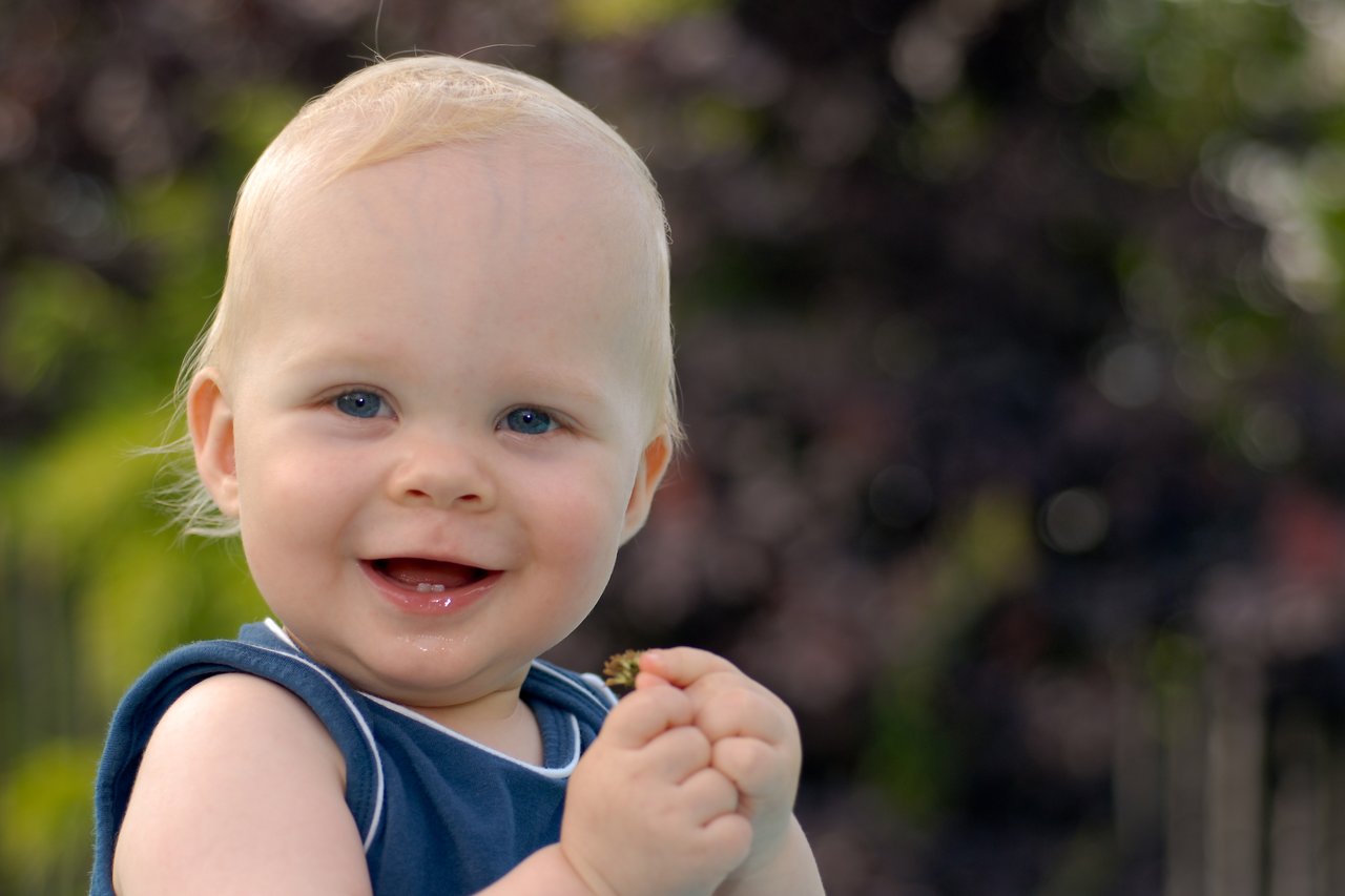 A smiling baby in a blue outfit holds a small object while looking at the camera outdoors.