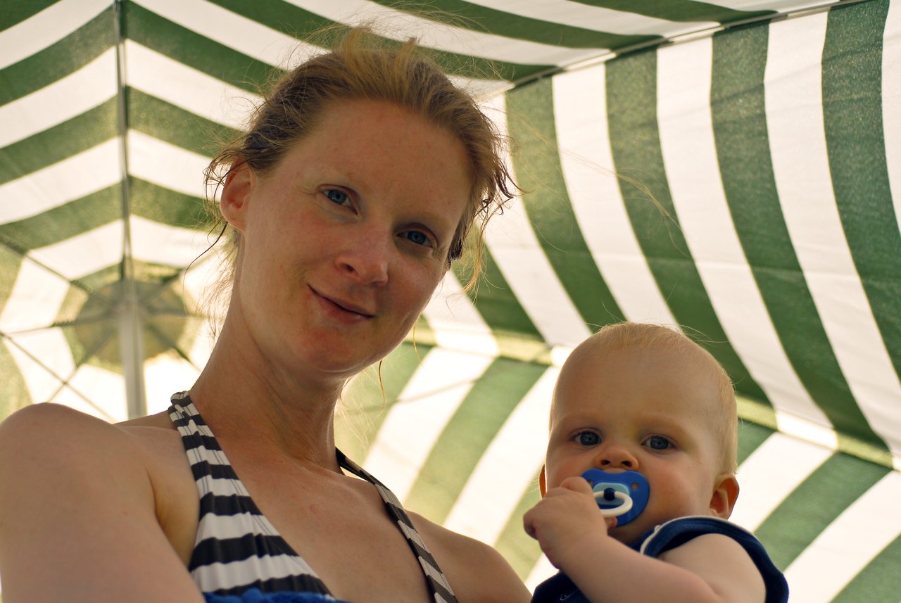 A woman in a striped swimsuit holds a baby with a pacifier under a green and white striped umbrella.