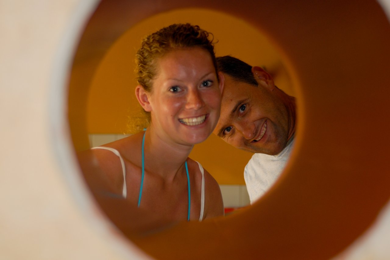Two people smiling and looking through a circular opening, with warm lighting in the background.