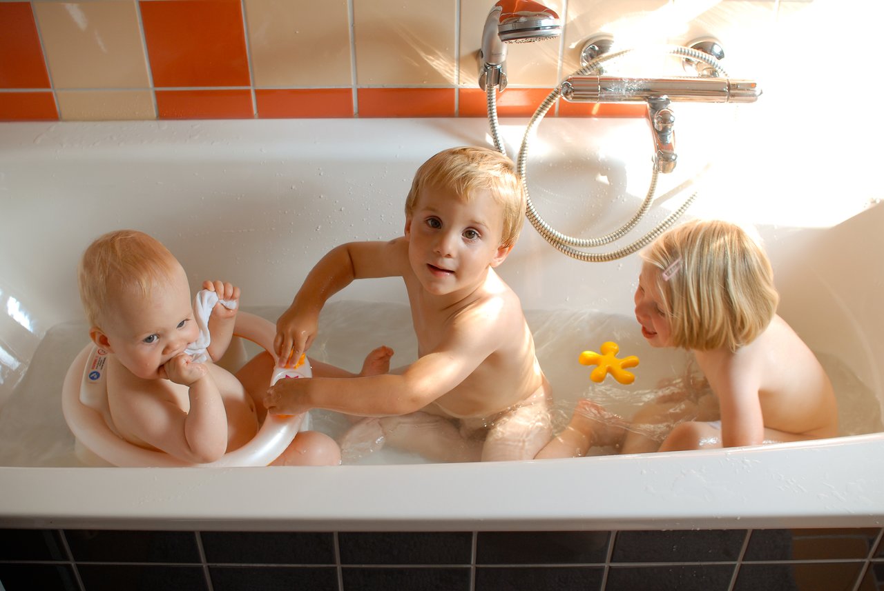 Three young children are playing together in a bathtub, splashing water and holding bath toys.