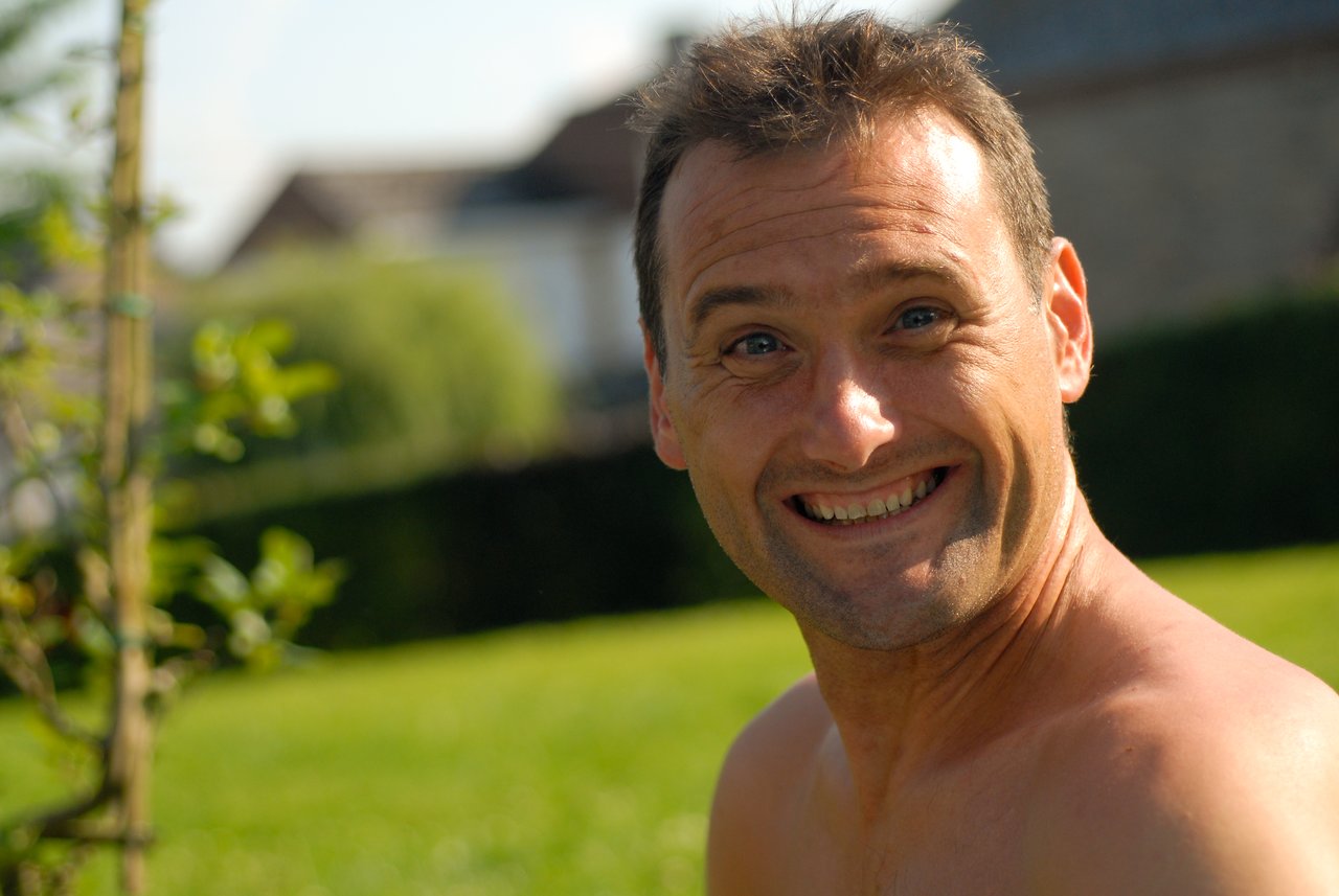 A smiling shirtless man looks at the camera outdoors with greenery and buildings in the background.
