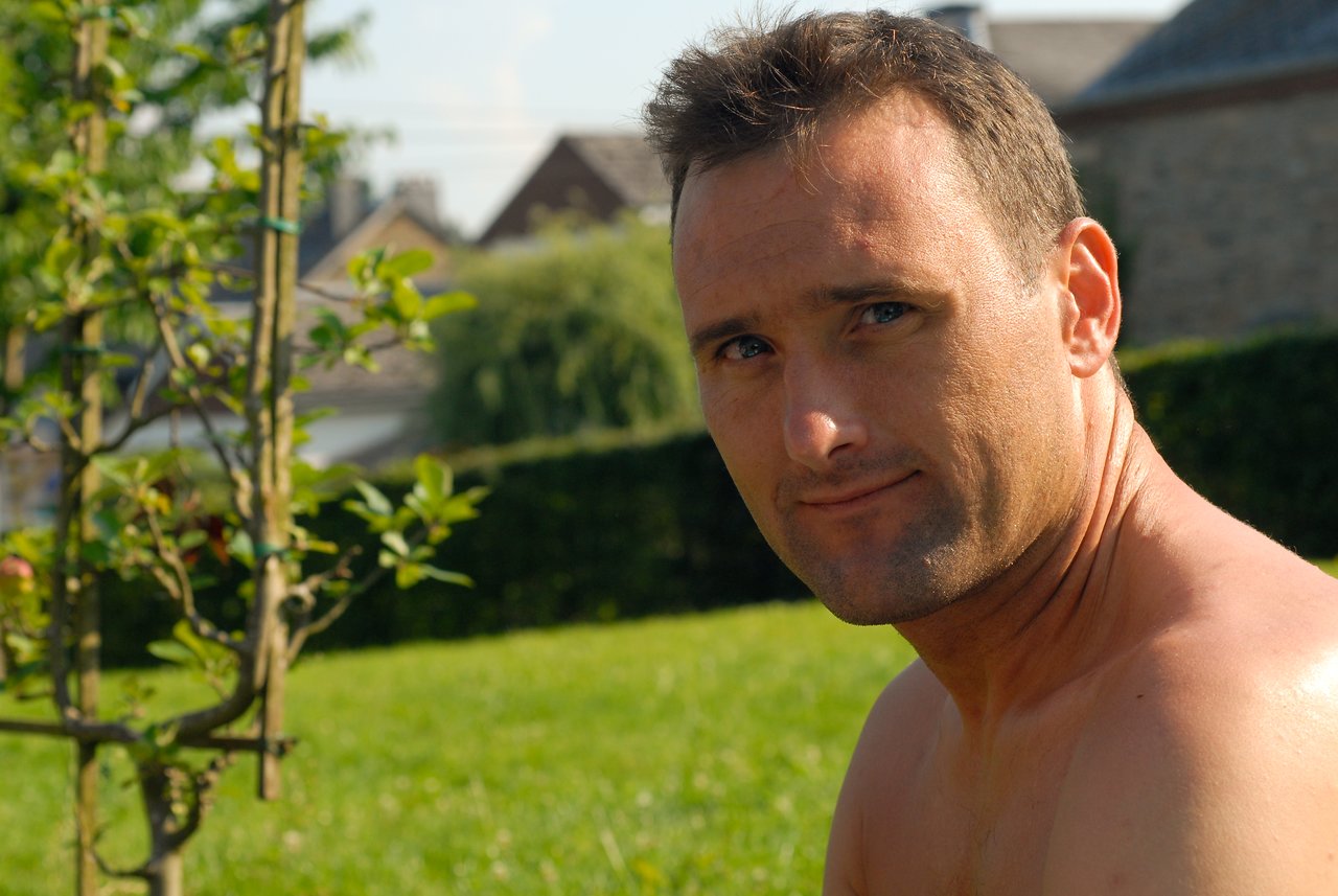 A shirtless man with short hair looks at the camera while sitting outdoors in a grassy area.