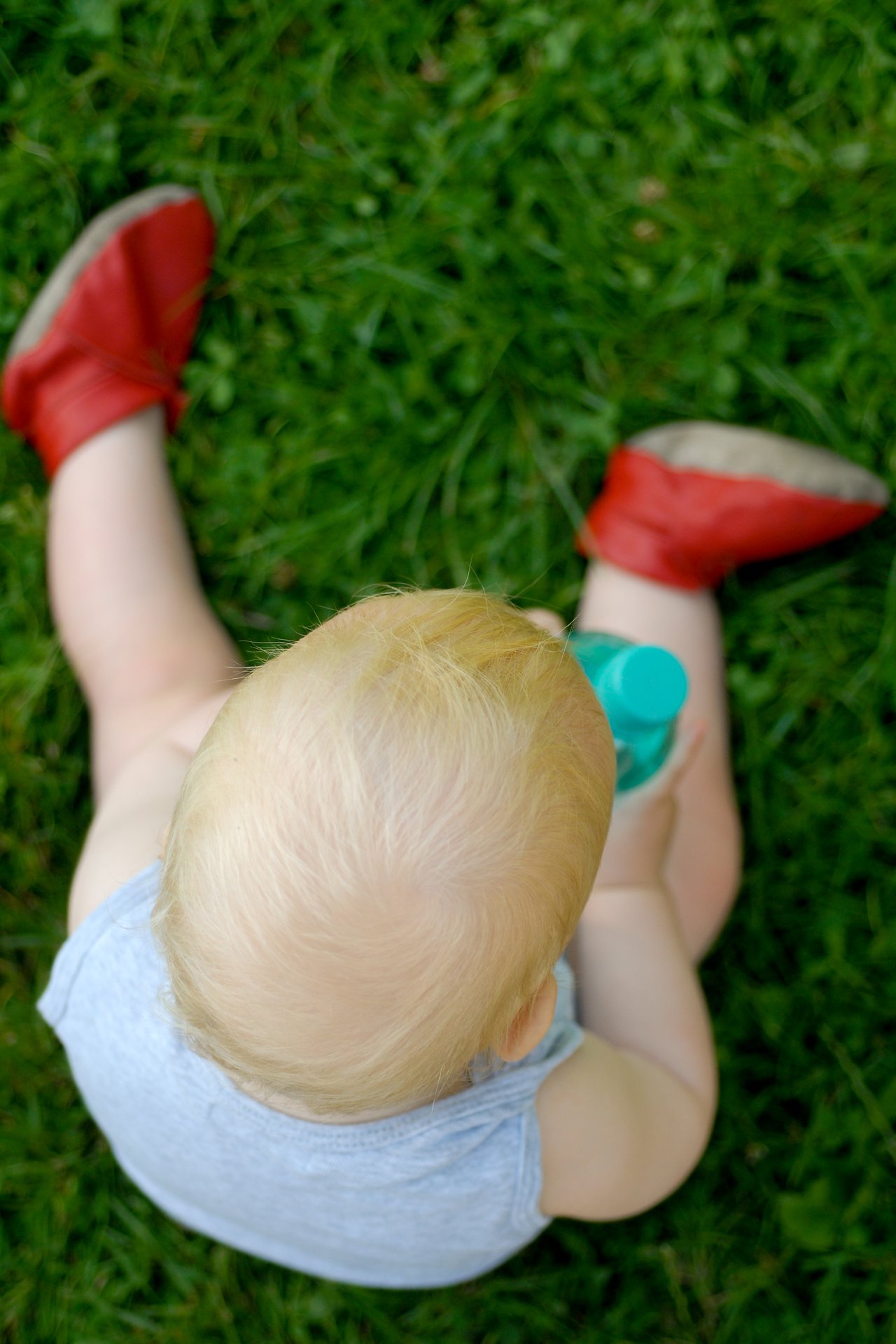 A baby with blonde hair sits on the grass, wearing red shoes and holding a small plastic bottle.