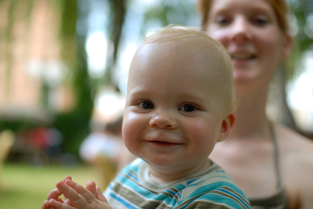 A smiling baby in a striped shirt claps hands, with a woman smiling in the blurred background.