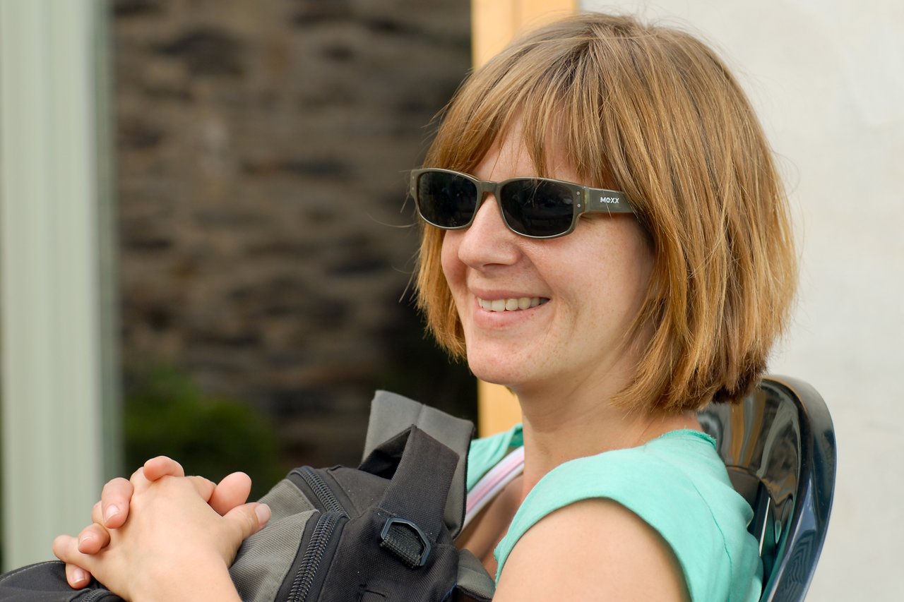A woman wearing sunglasses and a green top smiles while sitting and resting her hands on a backpack.