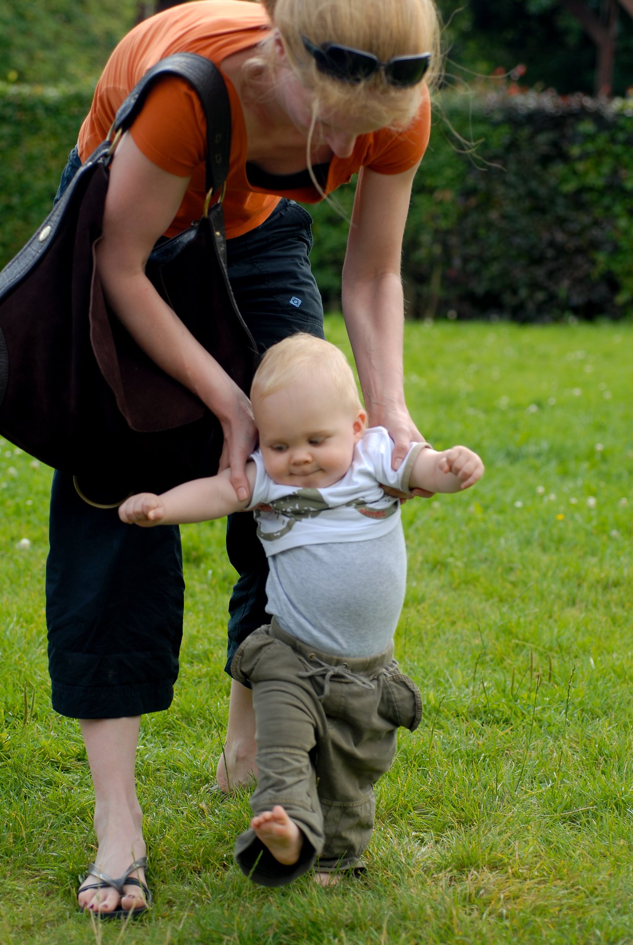 A woman helps a baby take steps on the grass, holding their arms for support.