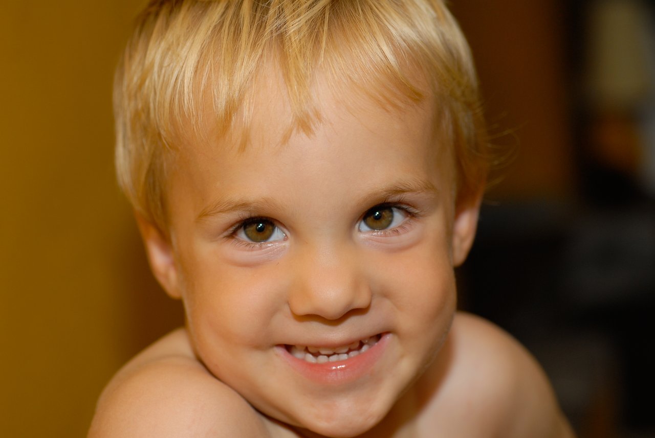 A young child with blonde hair smiles brightly while looking directly at the camera.