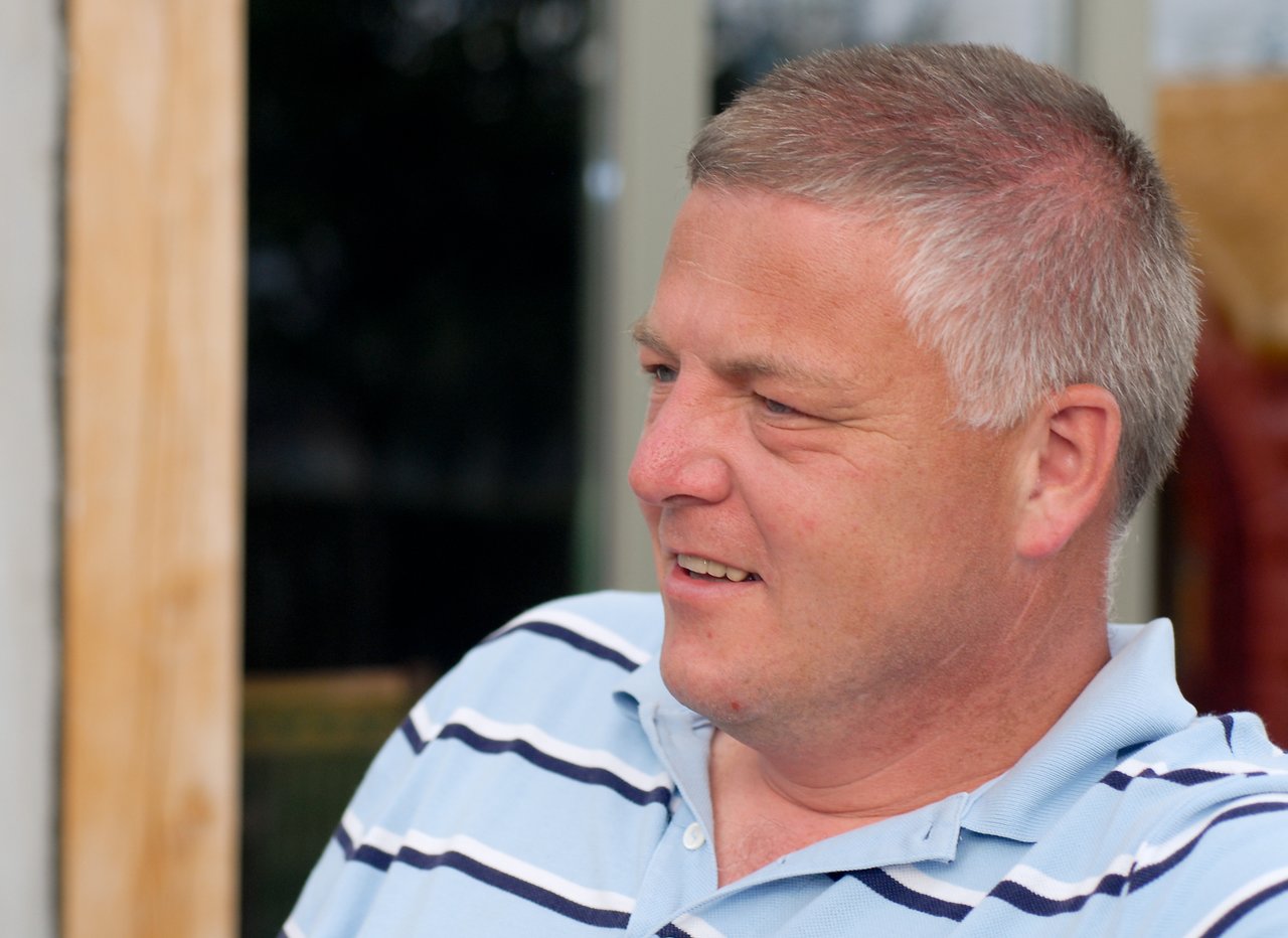 A man in a striped polo shirt sits outdoors, looking to the side with a slight smile.
