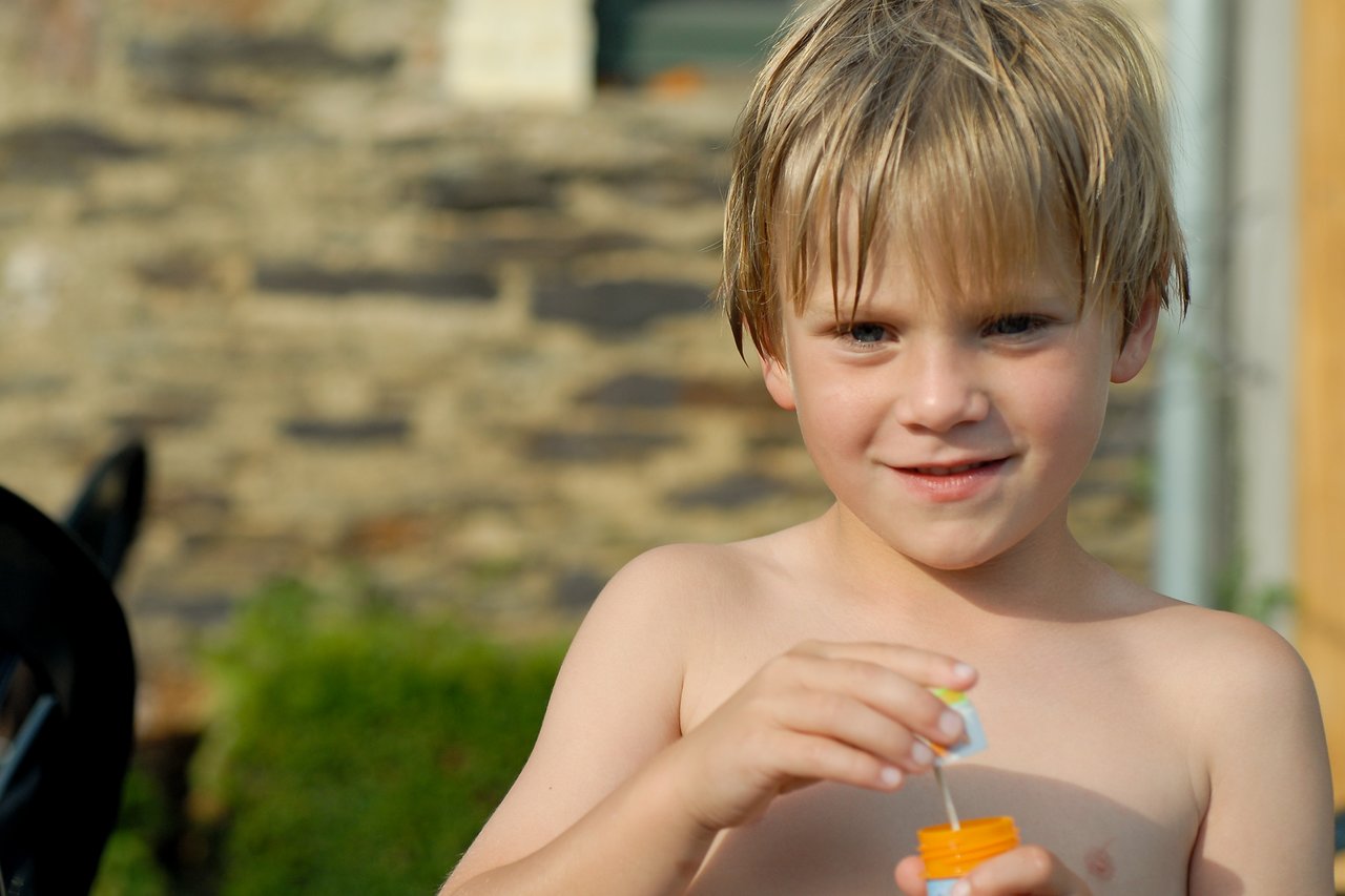 A shirtless child with wet hair holds a bubble wand and container, preparing to blow bubbles outdoors.