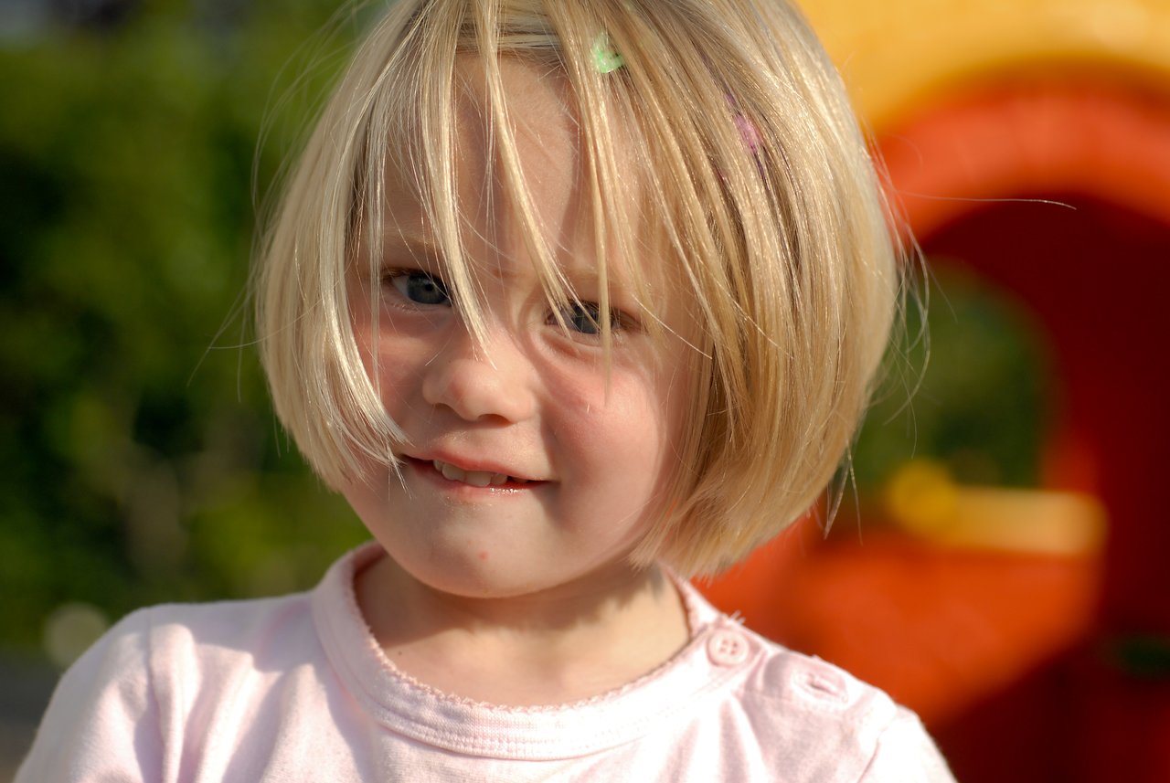 A young child with blonde hair and a pink shirt smiles slightly while standing outdoors in sunlight.
