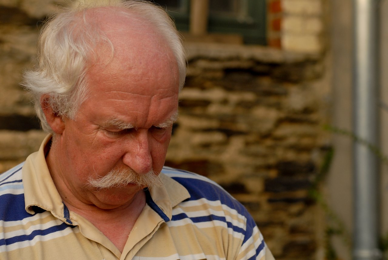An older man with white hair and a mustache looks down, wearing a striped polo shirt outdoors.
