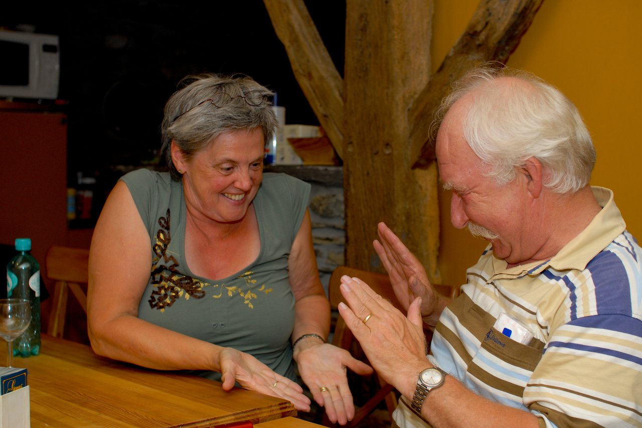 A woman and a man laugh while playing a hand game at a wooden table indoors.