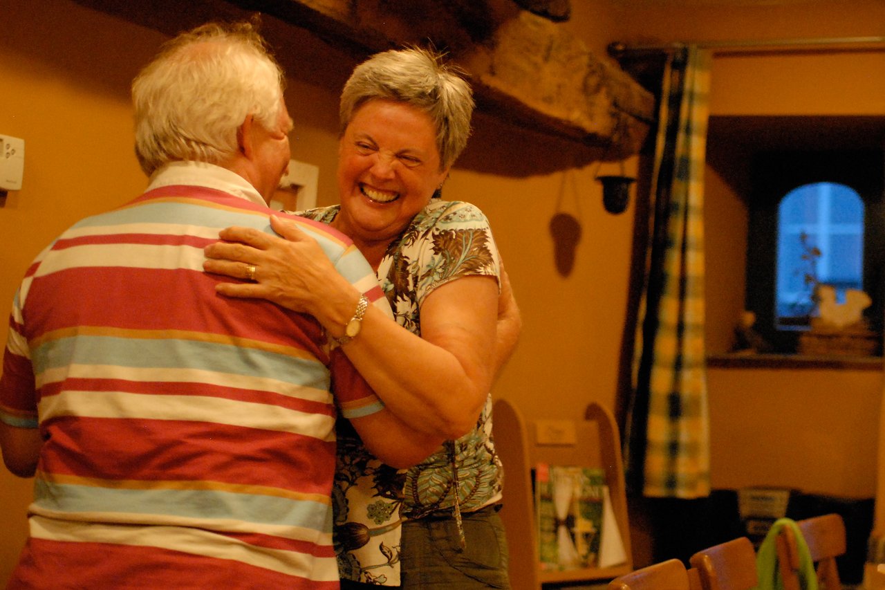 An elderly couple shares a joyful hug indoors, with the woman laughing and smiling warmly.
