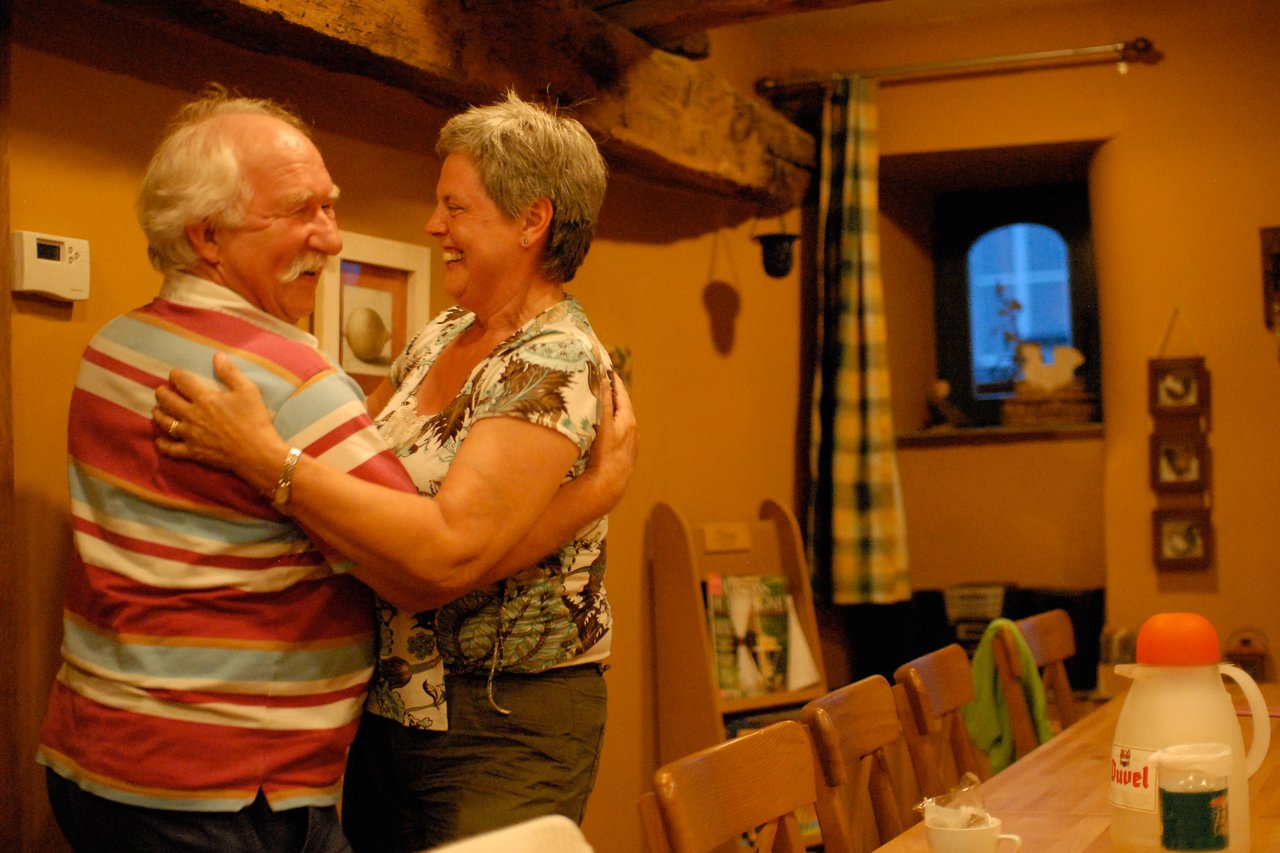 An elderly couple smiles and dances together in a warmly lit room, holding each other closely.