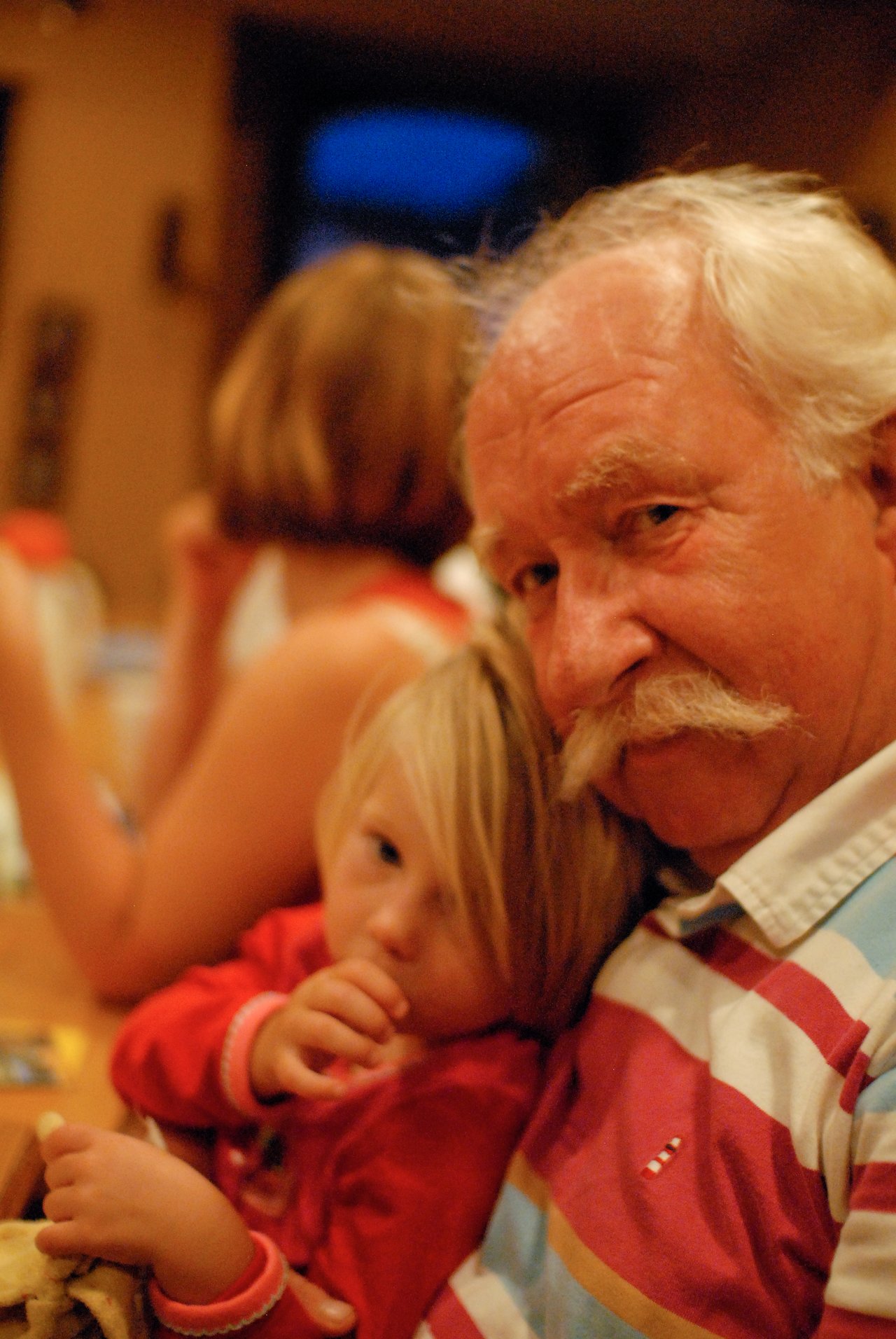 An older man with a mustache holds a young child in red, who is sucking their thumb and eating.