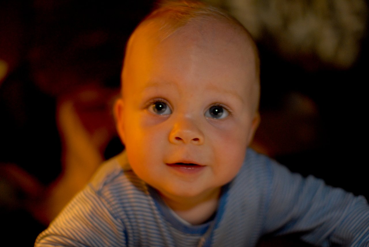 A baby in a striped shirt looks up with wide eyes and a slight smile in warm lighting.